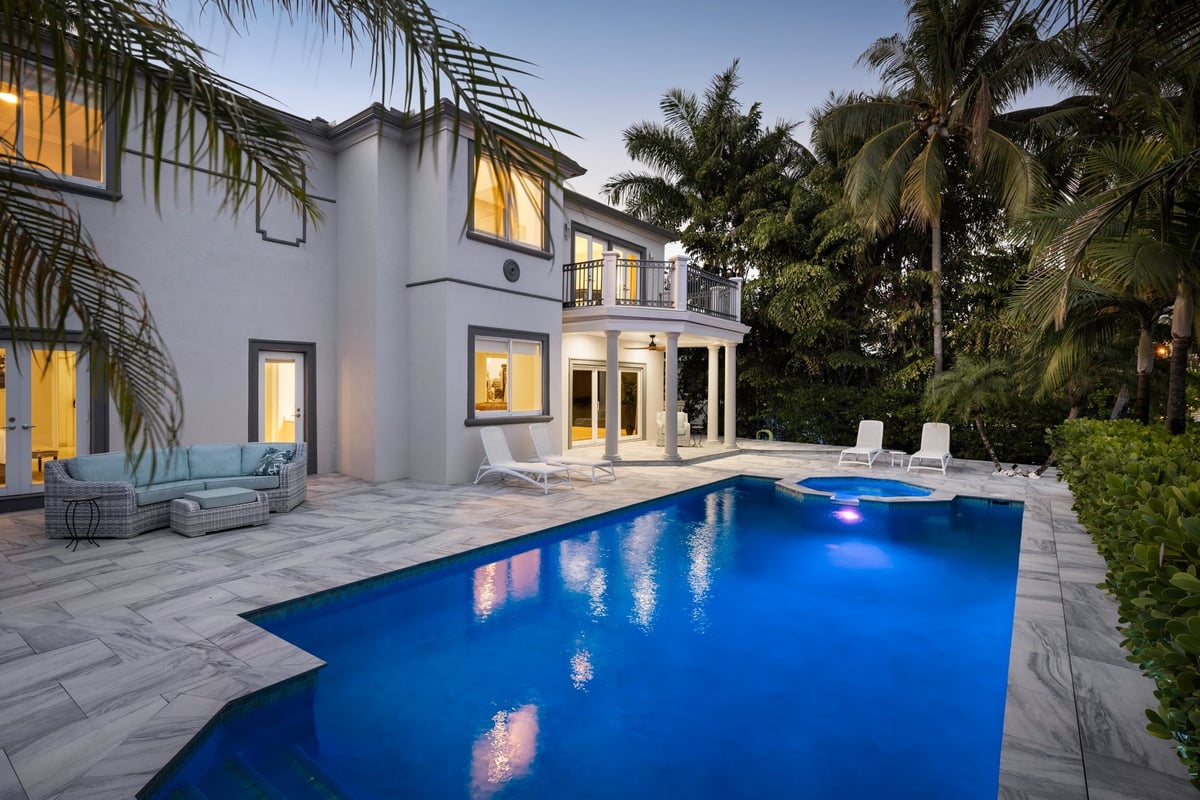 Backyard view of a stunning modern colonial-style house, part of a luxury residential neighborhood. Illuminated swimming pool surrounded by tropical palm trees and elegant landscaping.